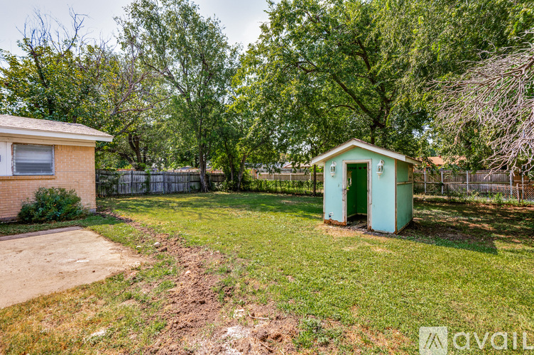 A small green shed sits in a yard with a brown house and trees in the background.