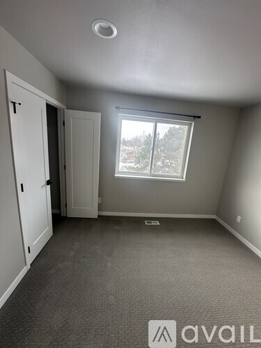 A room with a carpeted floor, a window with a view of trees outside, and a door on the left.