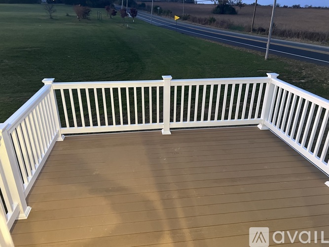 A wooden deck with white railings overlooks a grassy field and a road.