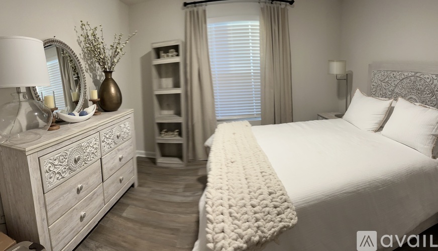 A well-lit bedroom with a large bed, a dresser with a mirror, and a nightstand with a lamp.