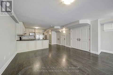 A spacious room with wooden flooring and white walls, featuring a kitchen area with white cabinets and appliances.