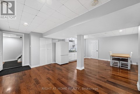 A spacious room with wooden floors and white walls, featuring a dining table and chairs, a kitchenette, and a staircase leading to another level.