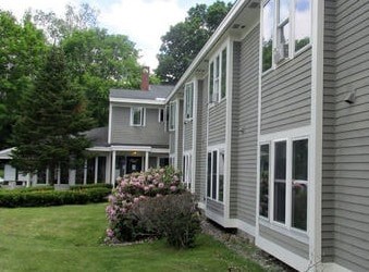 A grey house with a white window and a bush in front.