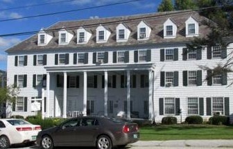 A white house with black shutters and a grey car parked in front.