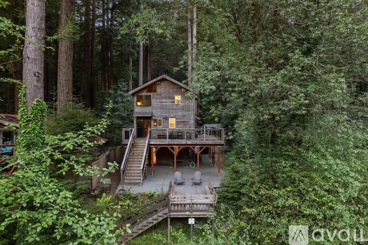 A wooden cabin is surrounded by trees and has a deck with chairs.