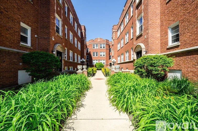 A pathway leads through a row of green plants in front of a brick building.