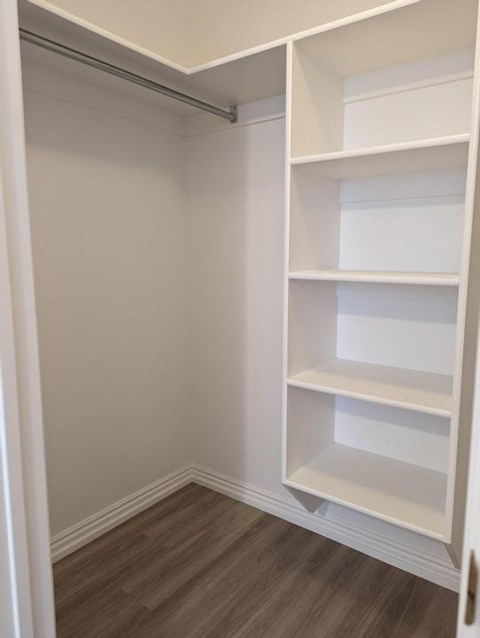 A white shelving unit with four shelves is open in a room with a white wall and wood flooring.