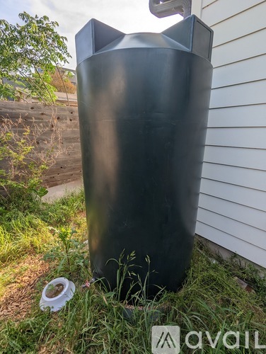 A tall black cylindrical object is placed in a grassy area next to a white siding wall.