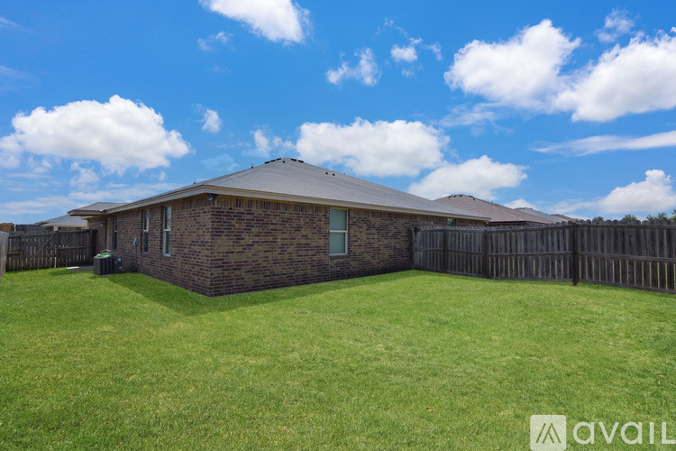A house with a brown brick exterior and a grey roof is surrounded by a green lawn and a wooden fence.