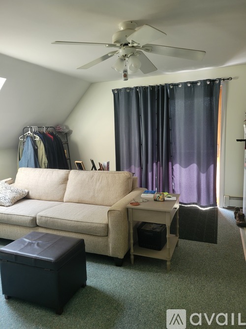 A living room with a beige couch, a black ottoman, and a ceiling fan.