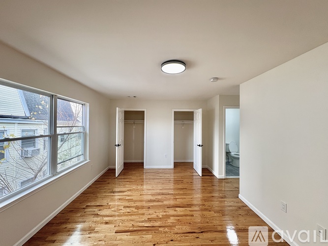 A spacious room with wooden floors and white walls, featuring a large window with a view of a tree-lined street.