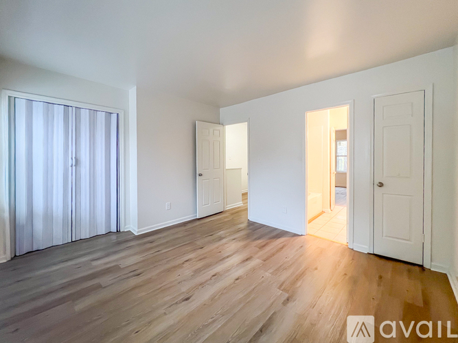 A room with wooden flooring and white walls, with a door on the right and a window with curtains on the left.