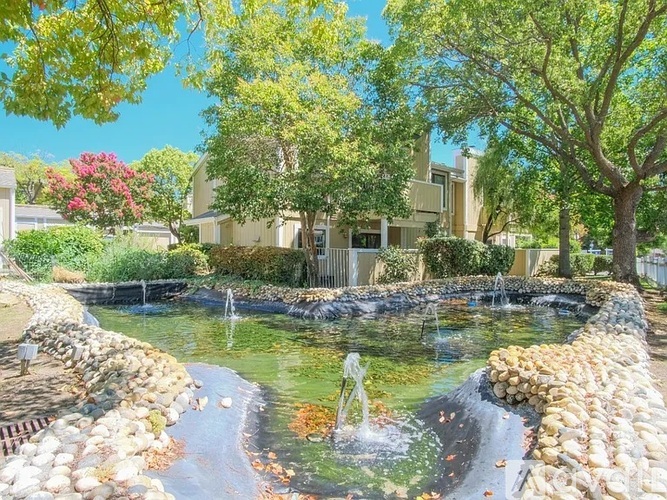 A pond with a waterfall and a fountain in the middle surrounded by rocks and trees.
