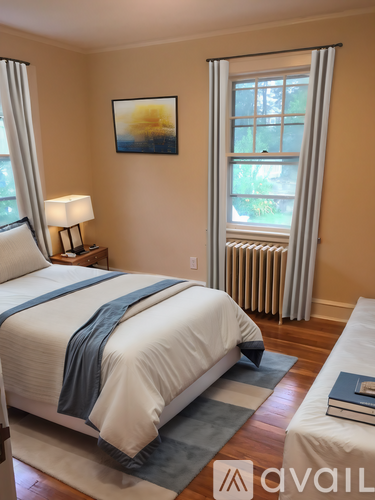 A bedroom with a bed, a nightstand with a lamp, a window with a view of trees, and a painting on the wall.
