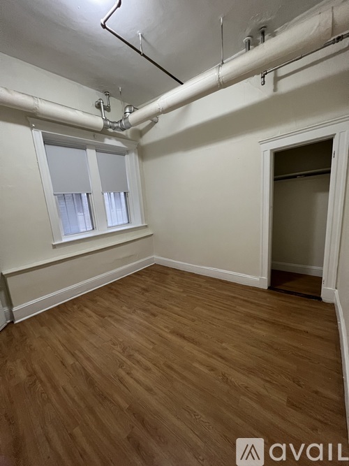 A room with wooden flooring and white walls, featuring a window with blinds and a door.