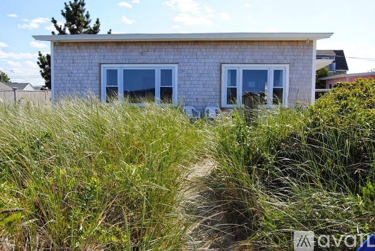 A house with a white brick exterior and two windows is surrounded by tall grass.