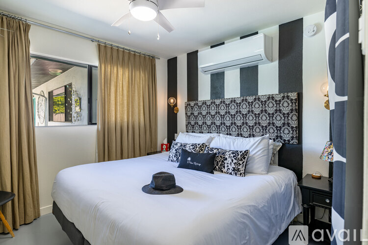 A bedroom with a bed, a hat on the bed, a window with curtains, a ceiling fan, and a wall with a black and white pattern.
