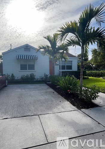 A house with a white exterior and a striped awning is surrounded by a palm tree and a concrete driveway.