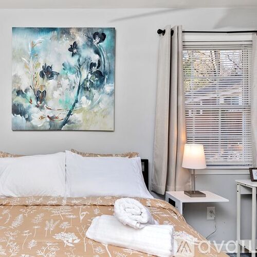 A bedroom with a large painting on the wall, a bed with a floral pattern, a small table with a lamp, and a window with blinds.
