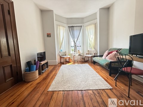 A living room with a large rug in the middle and a bed with a pink blanket in the background.