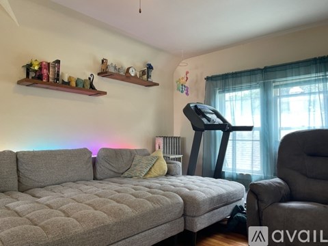 A living room with a grey couch, a grey ottoman, a grey chair, a grey wall, a grey window, a grey curtain, a grey floor, a grey wall, a grey wall, a grey wall, a grey wall
