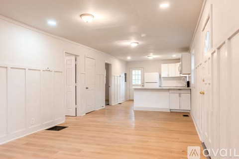 A spacious room with white walls and wooden flooring, featuring a kitchen area with white cabinets and a countertop.