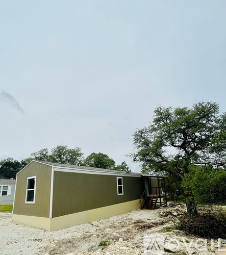 A small building with a green roof and tan walls sits on a gravel lot.