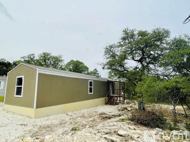 A small house with a brown roof and tan siding is surrounded by trees and gravel.