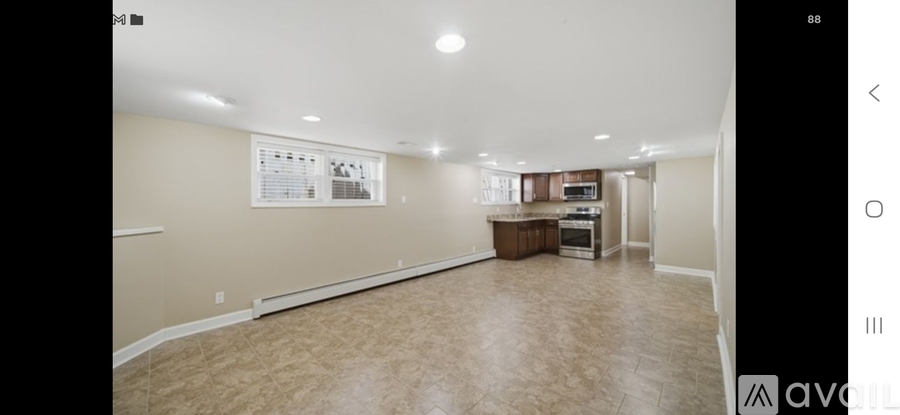 A spacious room with beige carpeting and a kitchen area in the background.