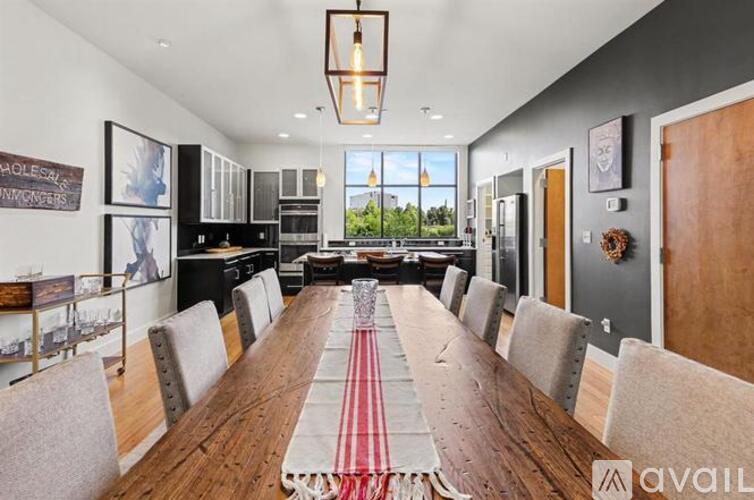 A long wooden table is surrounded by grey chairs in a room with a kitchen area in the background.