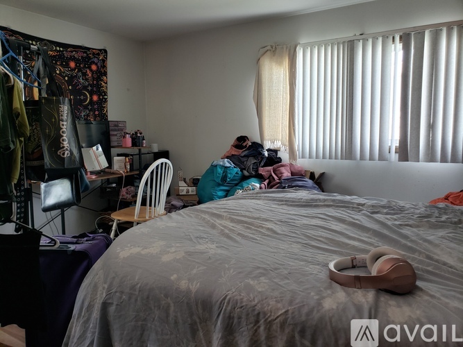 A messy bedroom with a bed in the center and a desk with a chair to the left.