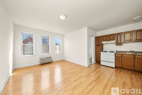 A room with wooden floors and white walls, with a window showing a view of a house, a radiator, and a kitchenette with a stove and cabinets.