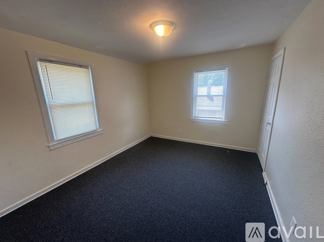 A room with a carpeted floor, a window with blinds, and a small window with a view of trees.