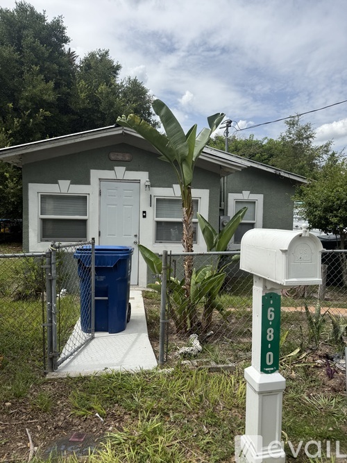 A small house with a grey roof and white door is surrounded by a fence and has a mailbox with the number 680 on it.