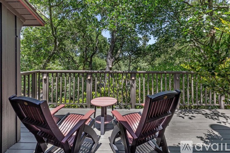 Two chairs and a table are on a deck with a railing and trees in the background.