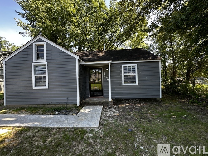 A small grey house with a black roof and a white door is surrounded by trees.