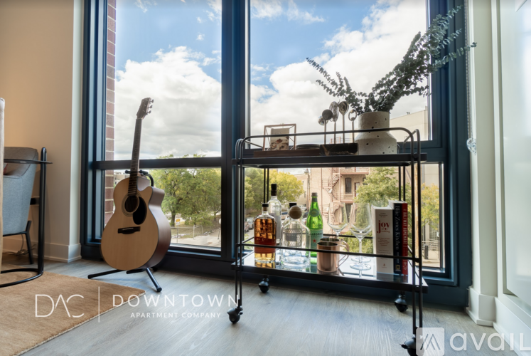 A room with a guitar, a shelf with bottles and a plant on it, and a window with a view of the sky and buildings outside.