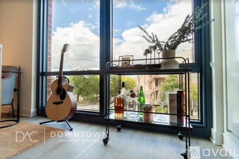 A room with a guitar, a shelf with bottles and a plant on it, and a window with a view of the sky and buildings outside.