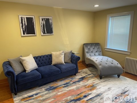 A blue couch with four pillows sits in a living room with a grey chair and two framed pictures on the wall.