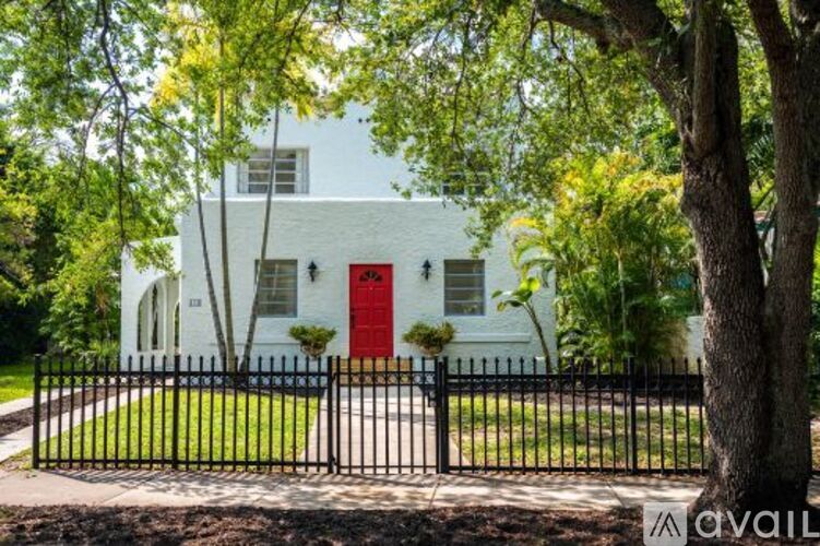 A white house with a red door is surrounded by a black fence and trees.