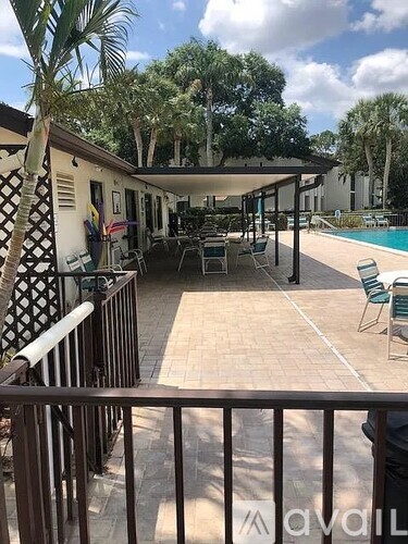 A patio with chairs and a table is surrounded by a pool and trees.