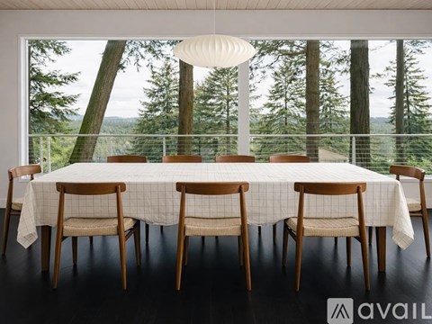 A table with chairs and a tablecloth in front of a window with a view of trees.
