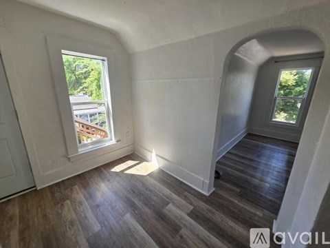A room with wooden flooring and white walls, featuring a large window and a doorway leading to another room.