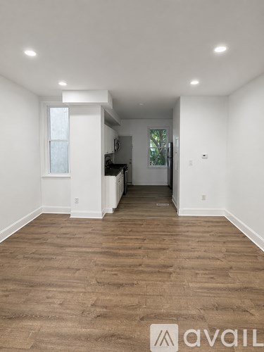 A room with wooden flooring and white walls, with a kitchen area visible in the background.