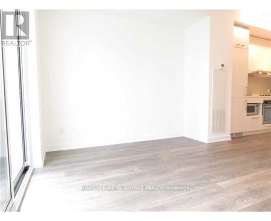 A large empty room with wooden flooring and a kitchen area in the background.
