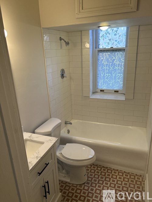 A white toilet sits in a bathroom with a white tub and a window.
