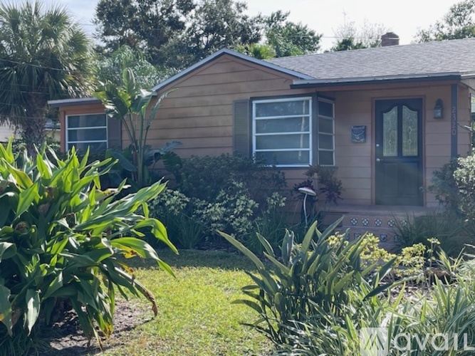 A house with a brown siding and a black door is surrounded by green plants.