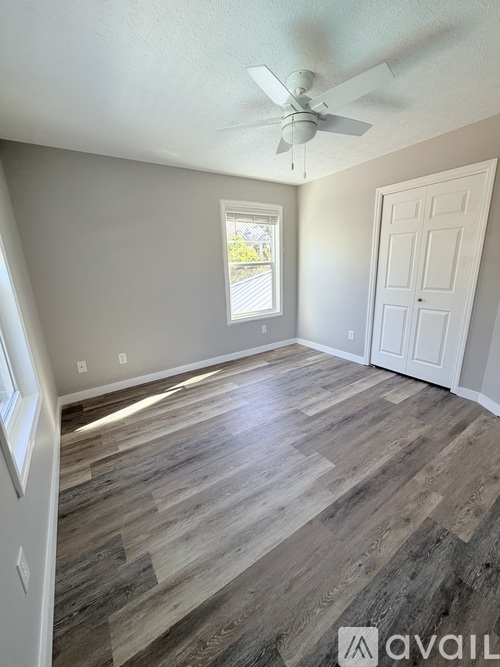 A room with a ceiling fan and light, hardwood floors, and a window with a view of trees outside.