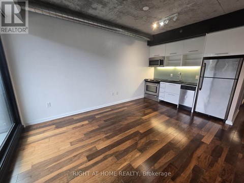 A room with wooden flooring and a kitchen area in the background.