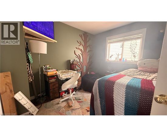 A bedroom with a bed covered in a multicolored blanket, a desk with a lamp, and a window with blinds.